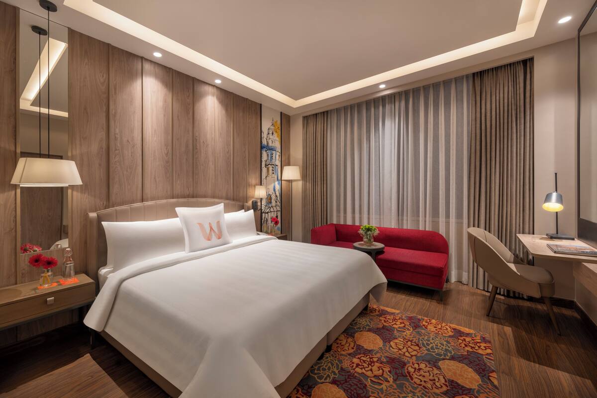 A modern hotel room with a large bed, a desk, a red armchair, and a patterned rug.