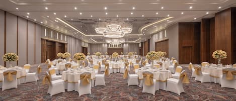 A banquet hall with round tables, white chairs, and gold sashes, featuring a patterned carpet and a chandelier.