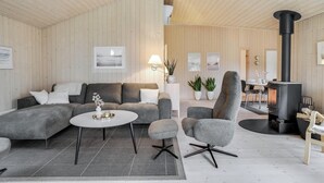 Un salon moderne avec un canapé d’angle gris, une table basse ronde et un fauteuil gris. On aperçoit un poêle à bois noir à l’arrière-plan.