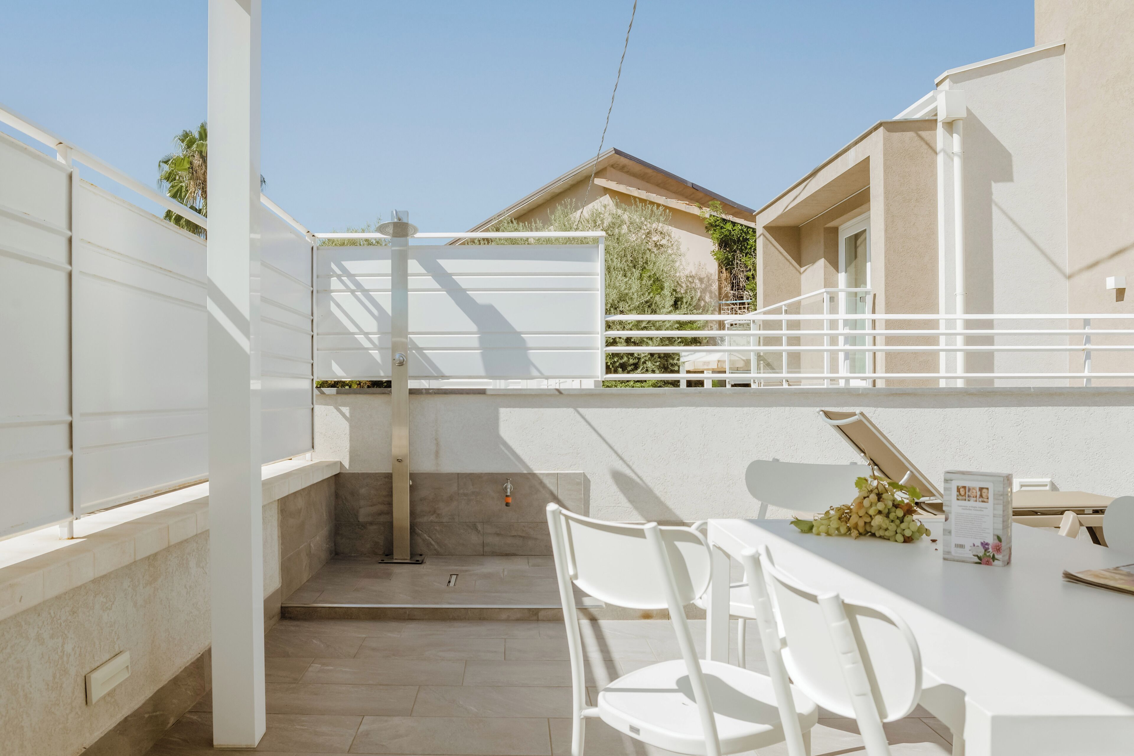 Un patio al aire libre con una mesa y sillas blancas, un pequeño jardín y vistas a un edificio con balcón.