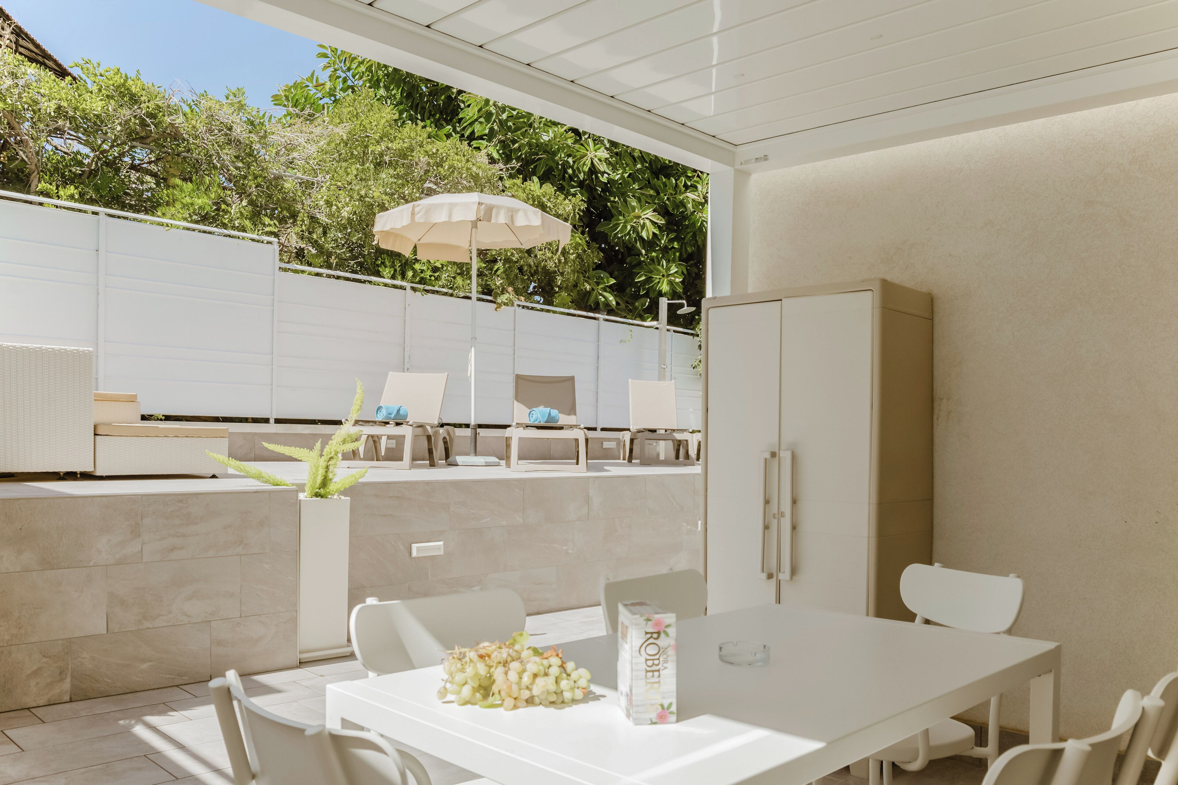 Eine Terrasse im Freien mit einem weißen Tisch, Stühlen und einem großen Sonnenschirm. Im Hintergrund befinden sich Kübelpflanzen und ein weißes Sideboard.
