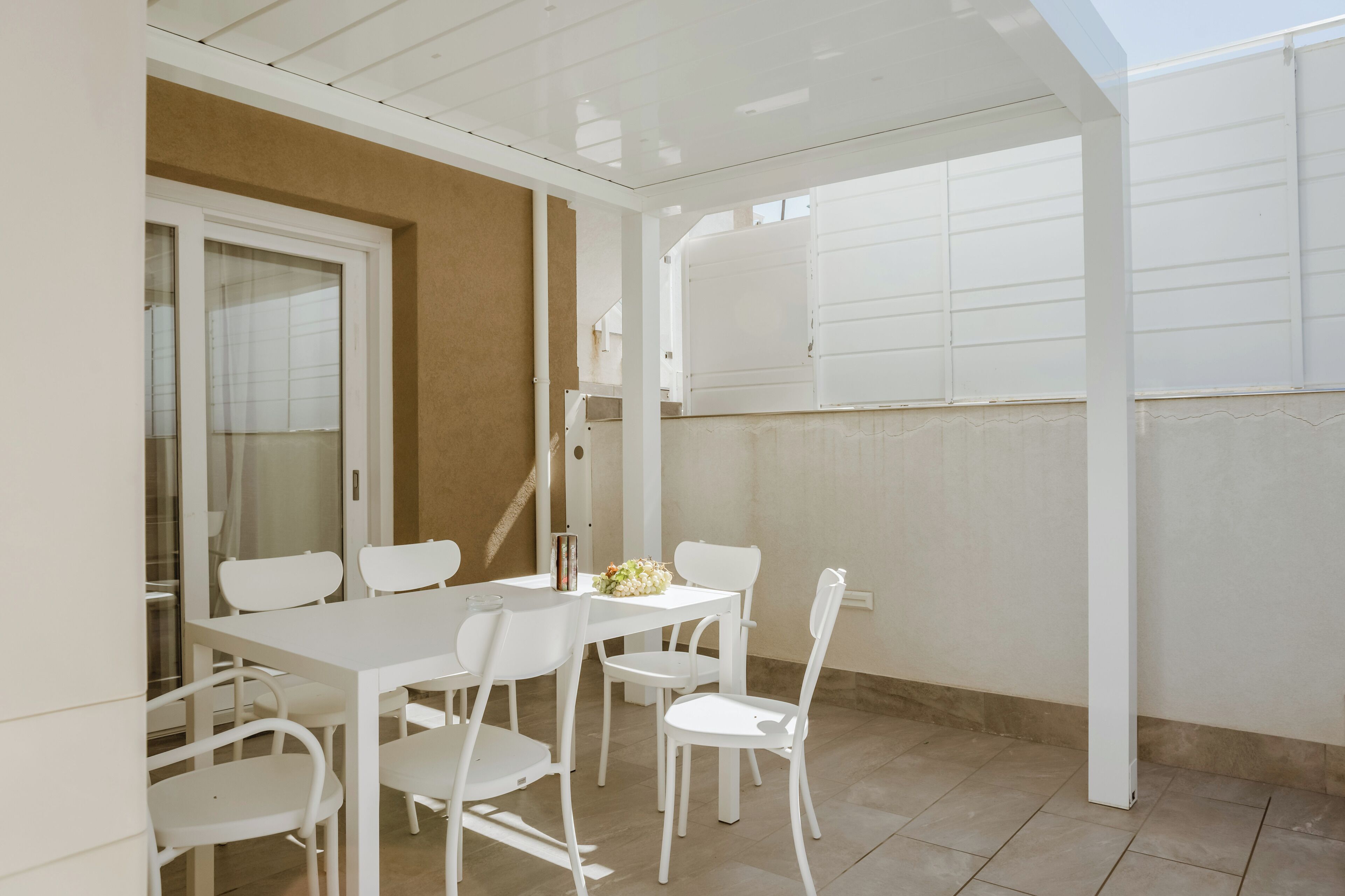 A white dining table with four chairs, a vase of flowers, and a window with blinds.