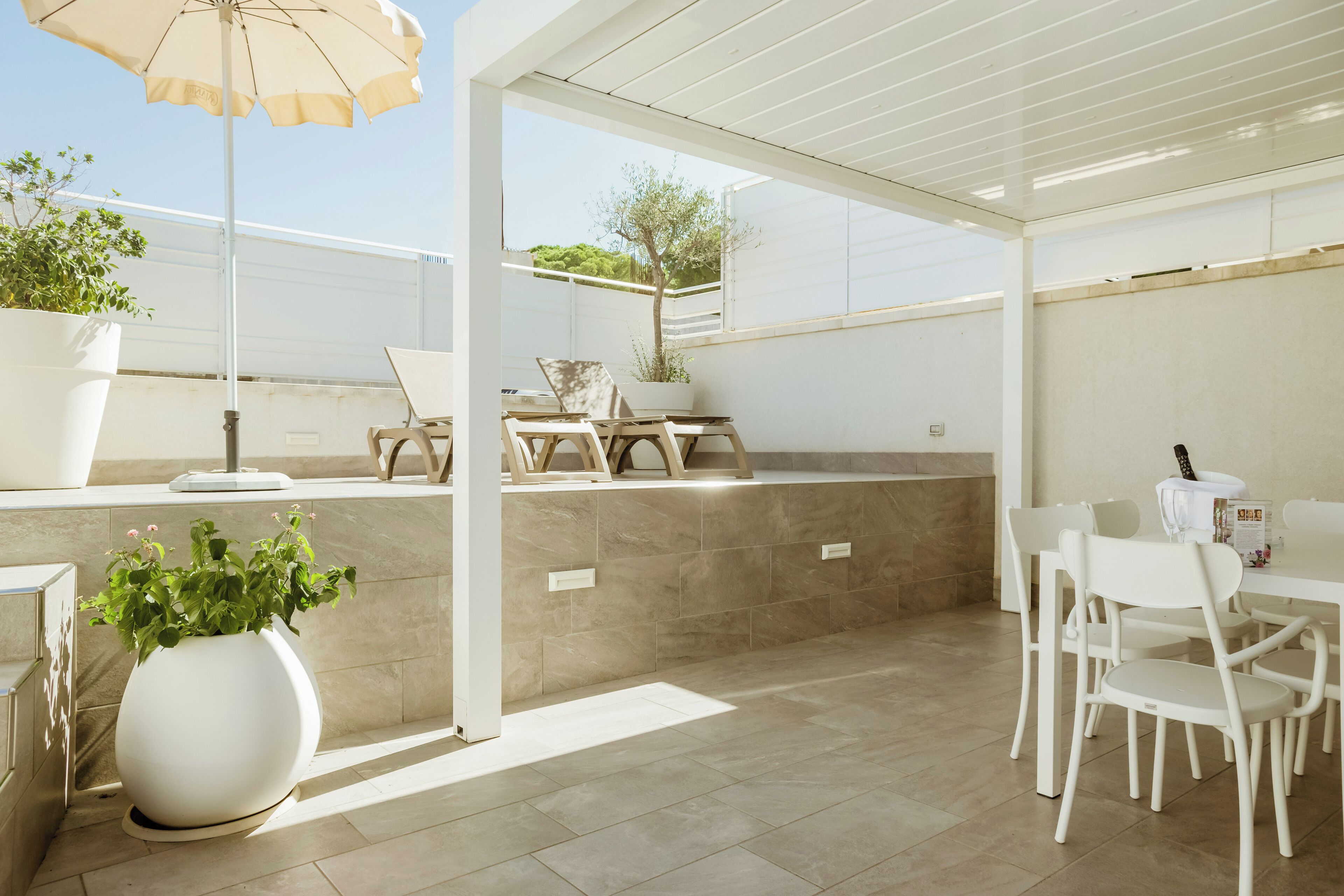A modern outdoor patio with a white table and chairs, a large potted plant, and a pergola with an umbrella.
