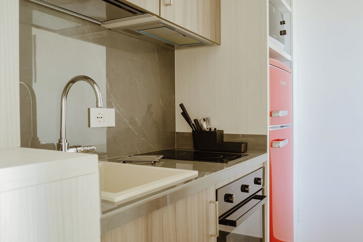 A kitchen with a modern design, featuring a stainless steel sink, a black induction cooktop, and a pink refrigerator.