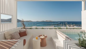 A modern balcony with a white interior, a seating area with cushions, a small round table with a drink, and a view of the sea and boats. - Aegean Colors Paros (Paros)
