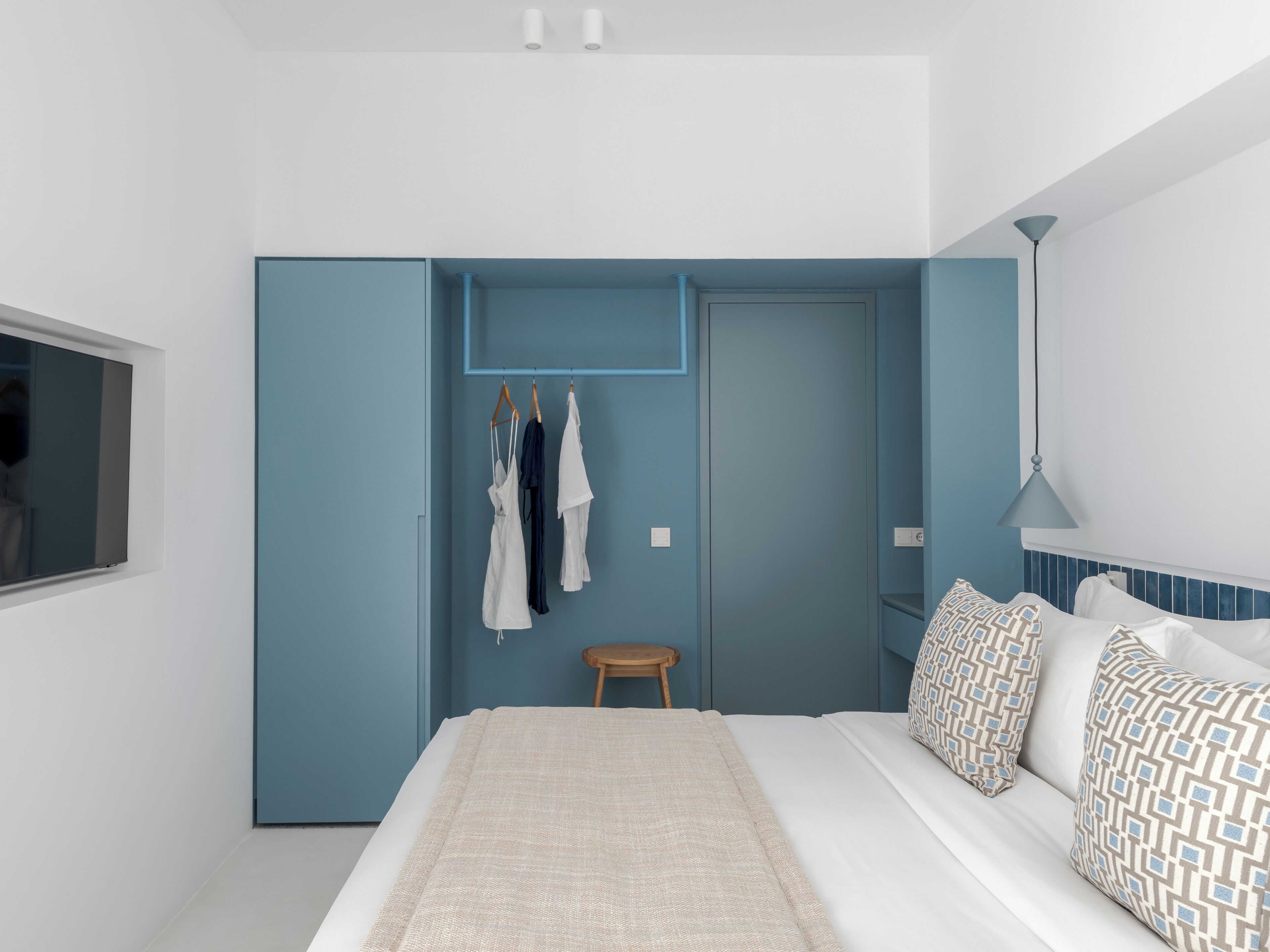 Une chambre à coucher comprenant un lit, une armoire, un portant à vêtements, un petit tabouret et une lampe.