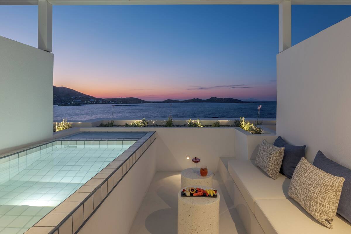A modern outdoor seating area with a pool, overlooking a coastal view at sunset.
