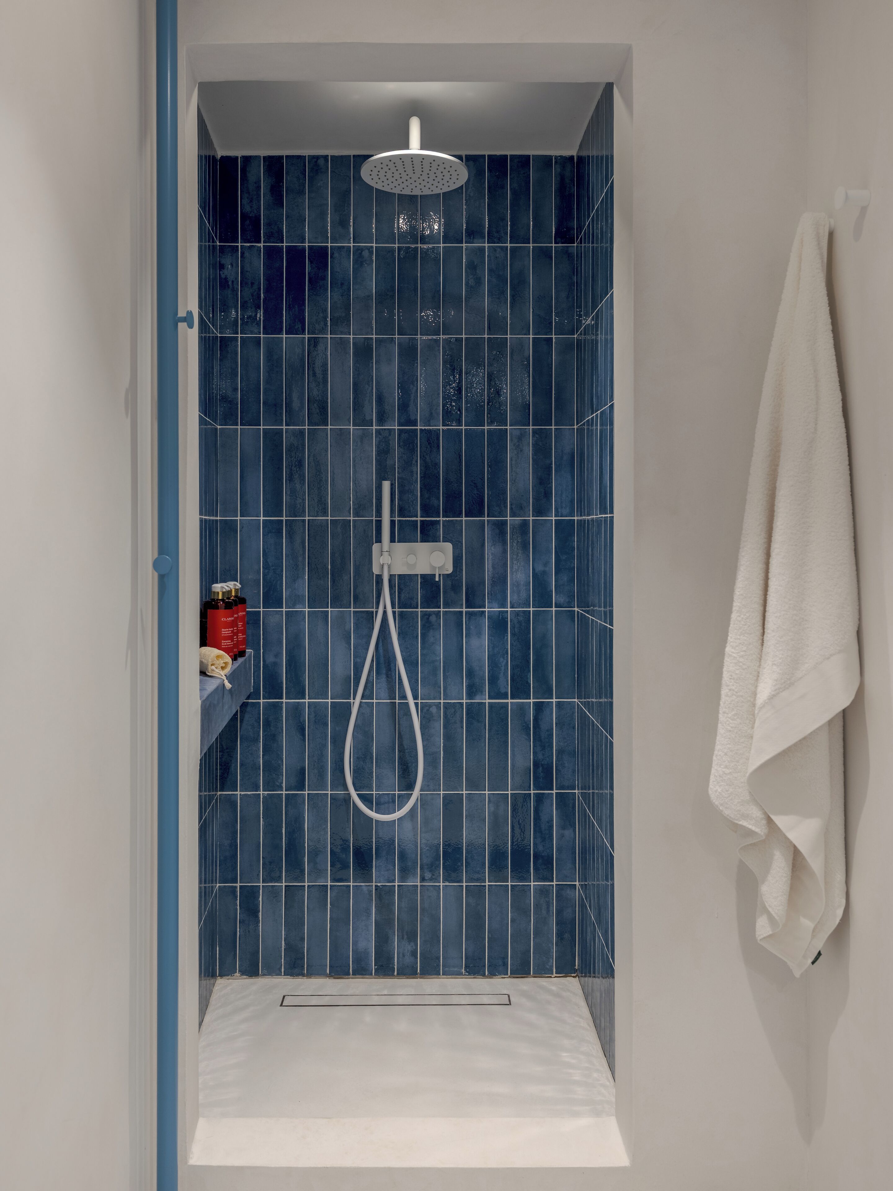 A shower with a handheld showerhead, a white towel hanging on the wall, and a shelf with toiletries.