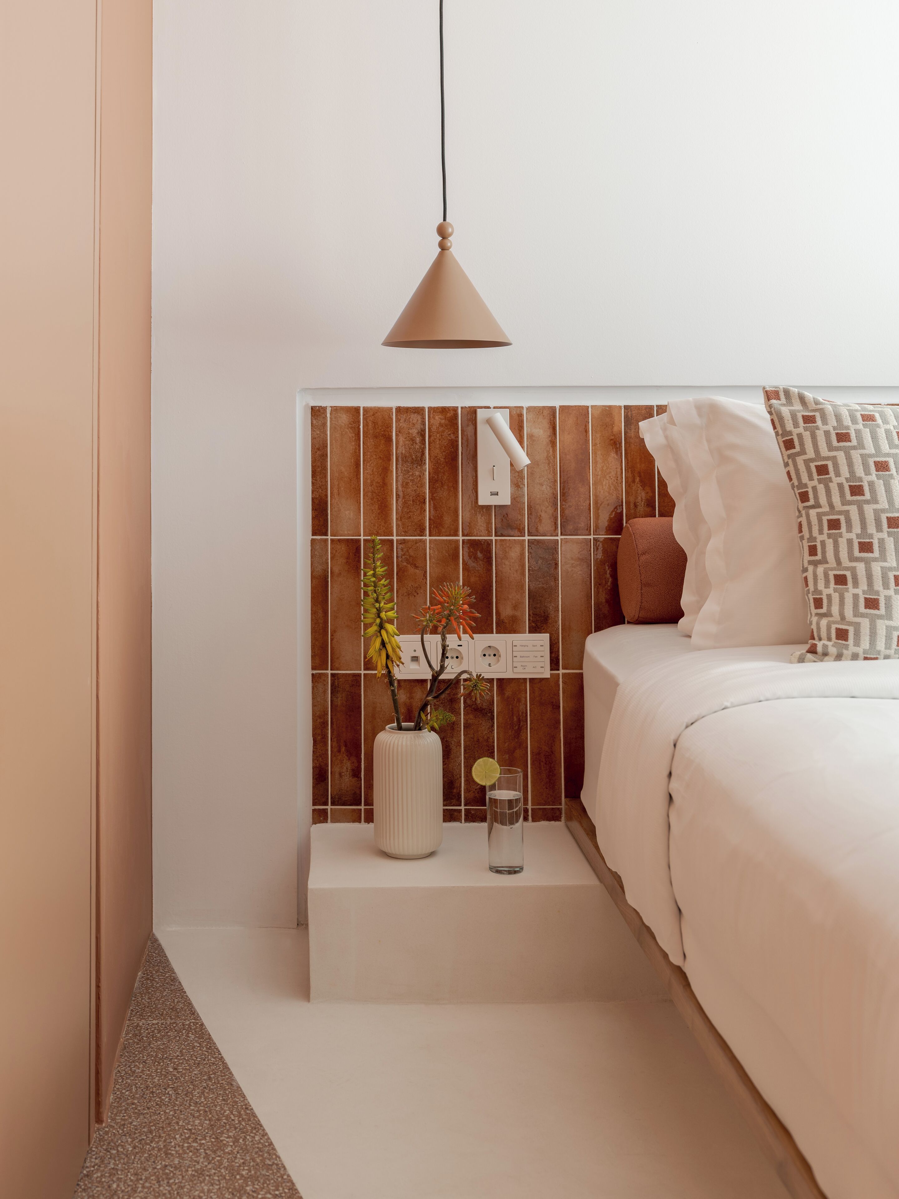 A bedroom with a bed, a bedside table with a vase and a glass, and a wall-mounted light fixture.