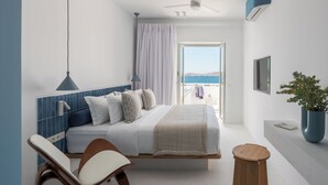 A modern bedroom with a bed, a bedside chair, a small wooden stool, a potted plant, and a view of the sea through an open door. - Aegean Colors Paros (Paros)
