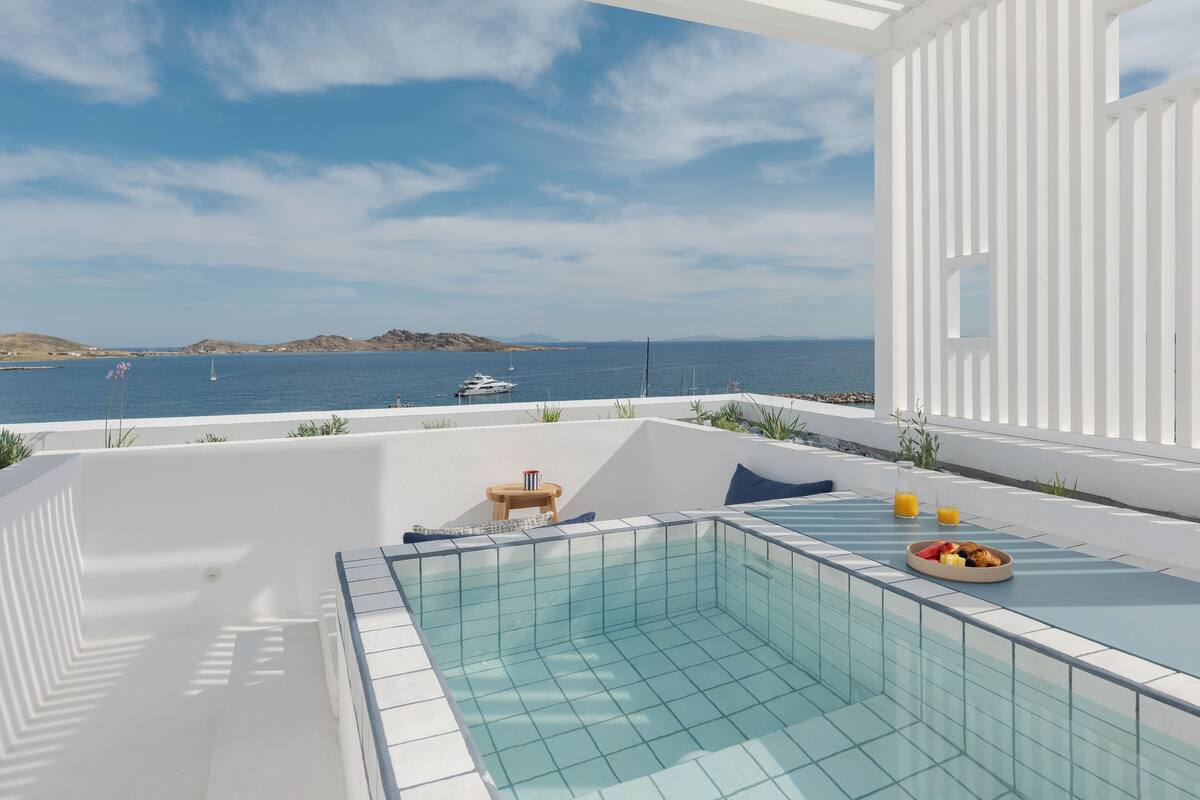 A pool area with a view of the sea and boats, a white exterior wall, and a small seating area with a table and cushions.