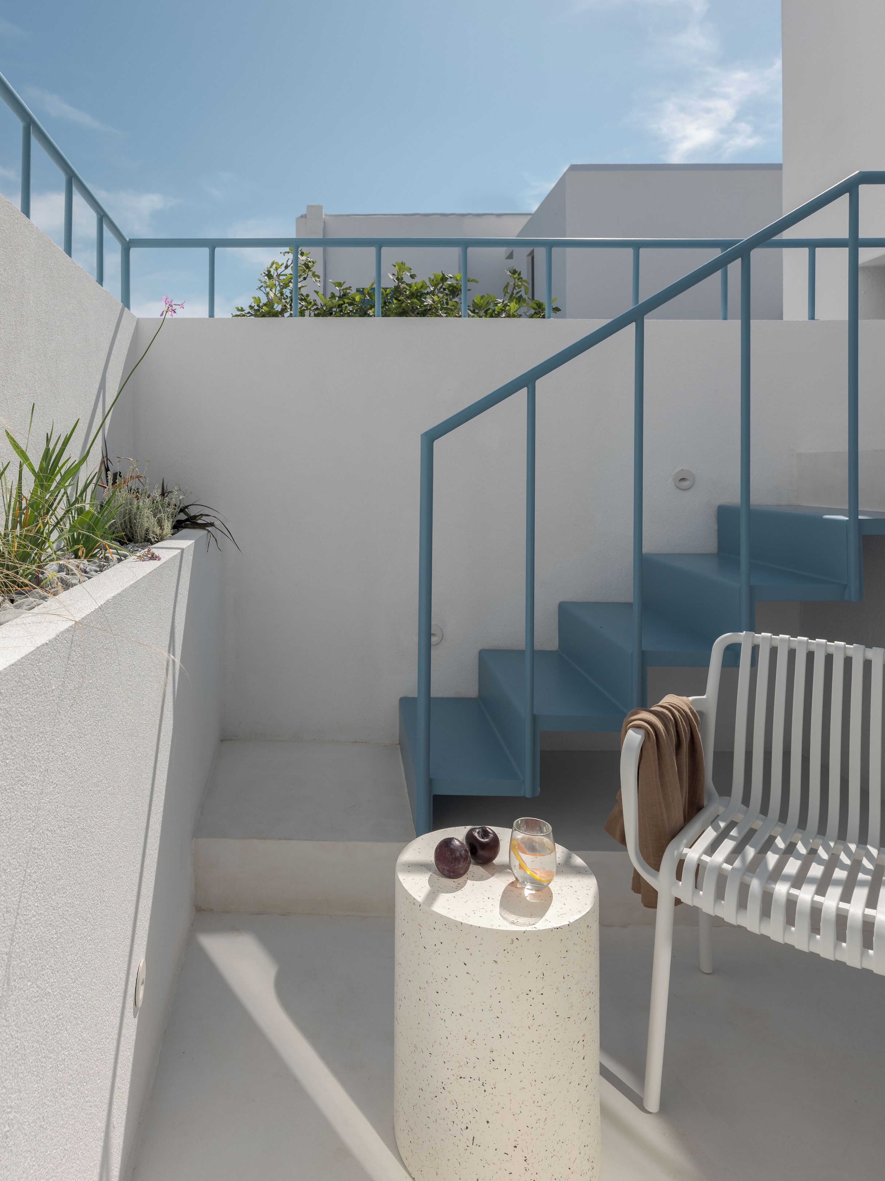 Un área de estar exterior moderna con un banco blanco, una pequeña mesa redonda con una bebida y fruta, y una escalera azul.