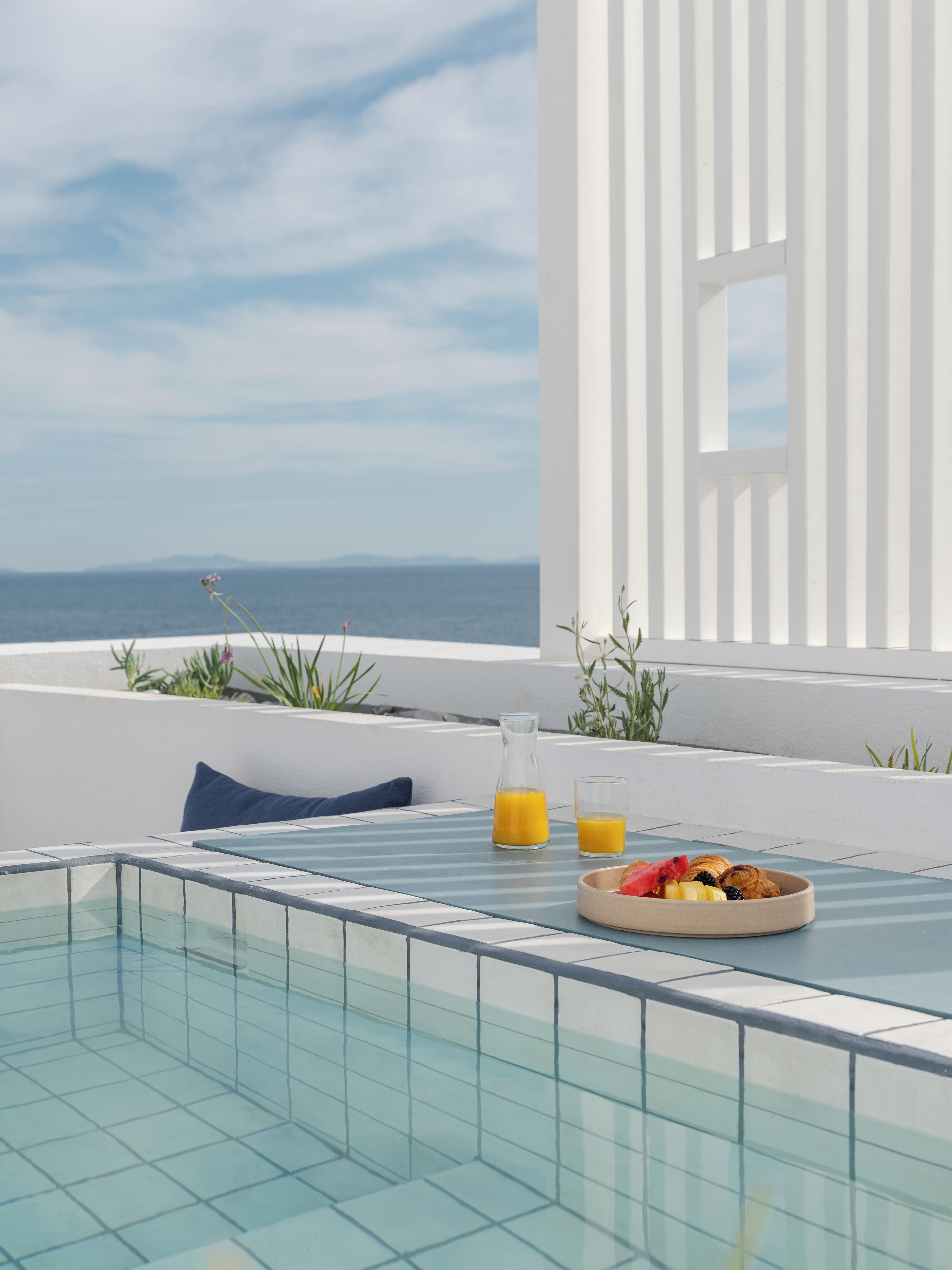 Zona junto a la piscina con una mesa, un bol de fruta y dos vasos de zumo de naranja.