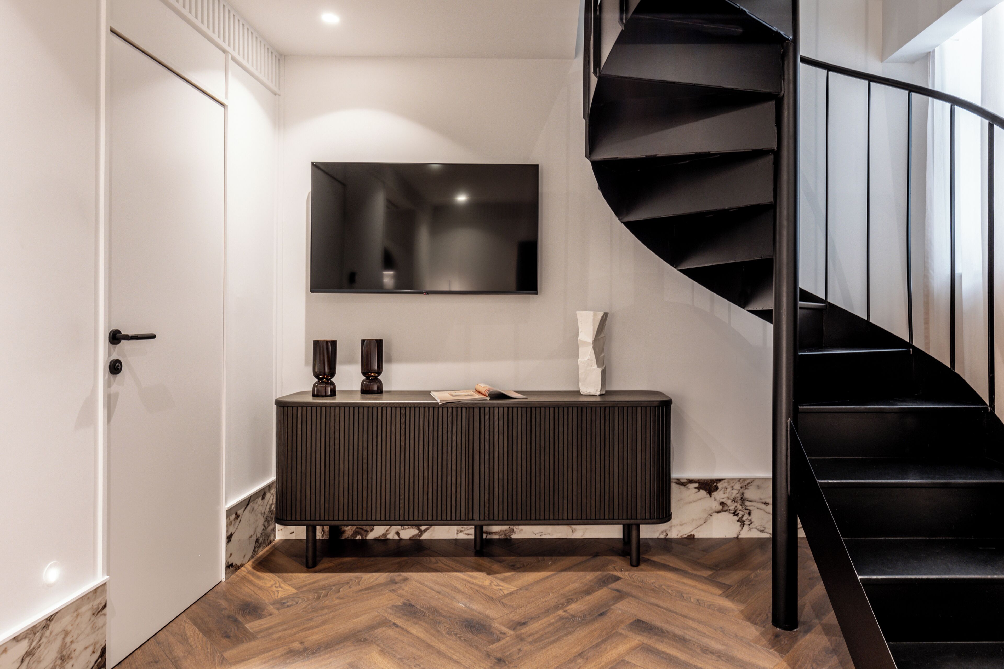 Un interior moderno con una escalera espiral negra, un televisor de pantalla plana montado en la pared y un mueble consola de madera oscura.