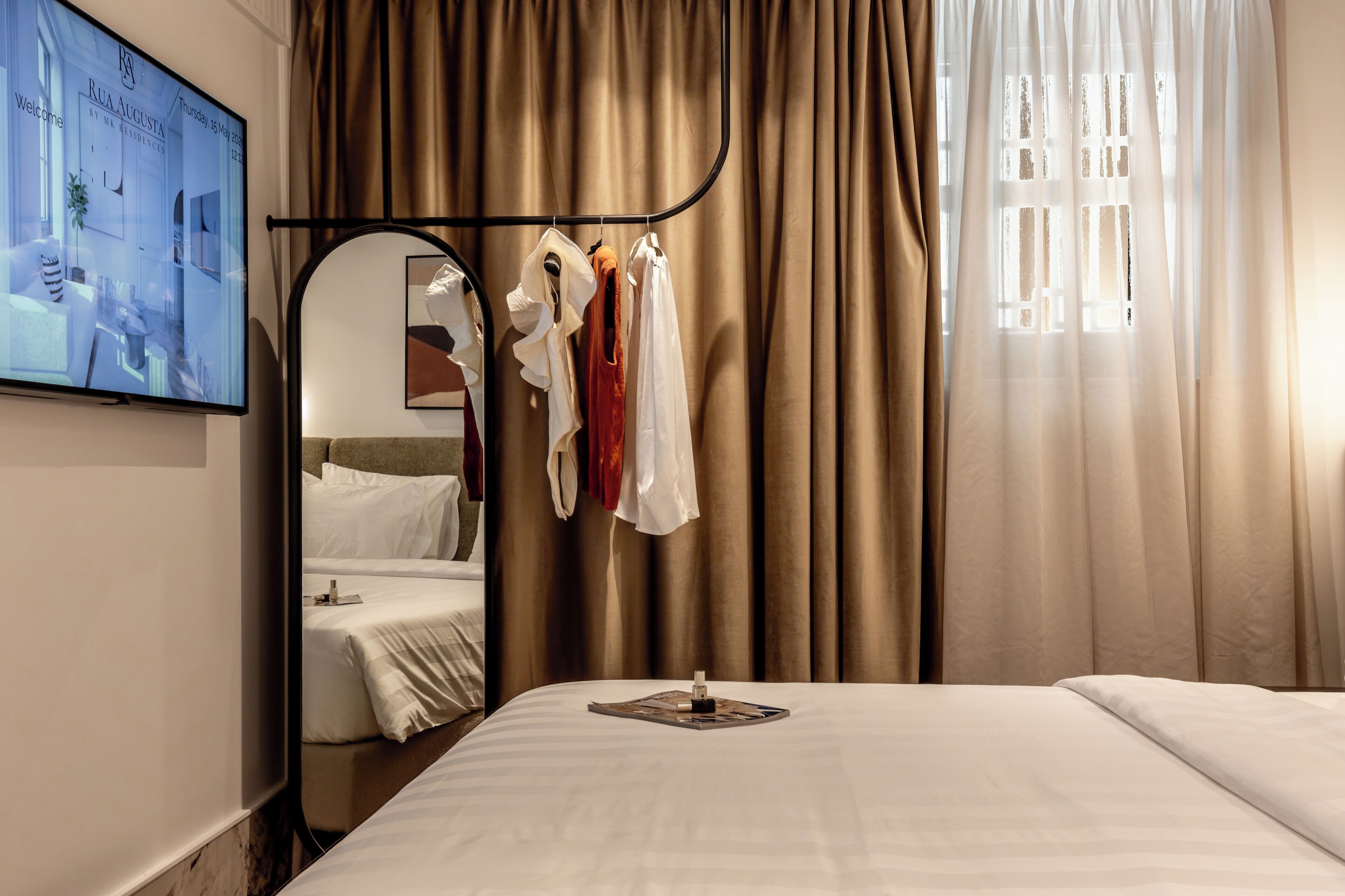 A hotel room with a bed, a TV mounted on the wall, a mirror, and a clothing rack.