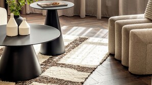 Un salon moderne avec une table basse ronde noire, un canapé beige et une grande fenêtre ornée de rideaux légers.