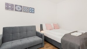 A modern room with a grey sofa, a bed with pink and white pillows, and a grey blanket. There are three patterned wall hangings above the sofa. - TOTU Home Gdańsk Marina Studio Old Town (Gdansk)