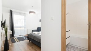 A modern bedroom with a bed, bedside table, and a view of the city through a large window. - TOTU Home Gdańsk Marina Studio Old Town (Gdansk)