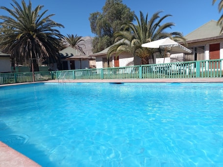 A clear blue pool with a green fence, palm trees, and buildings in the background.. Cabañas Yunkai