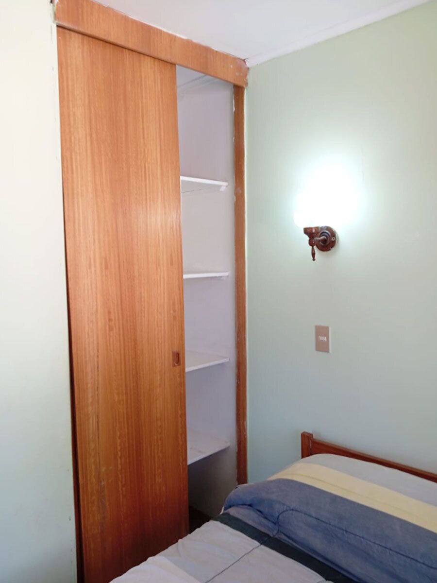 Una habitación con un armario de madera, una cama con colcha a rayas y una lámpara fijada a la pared.