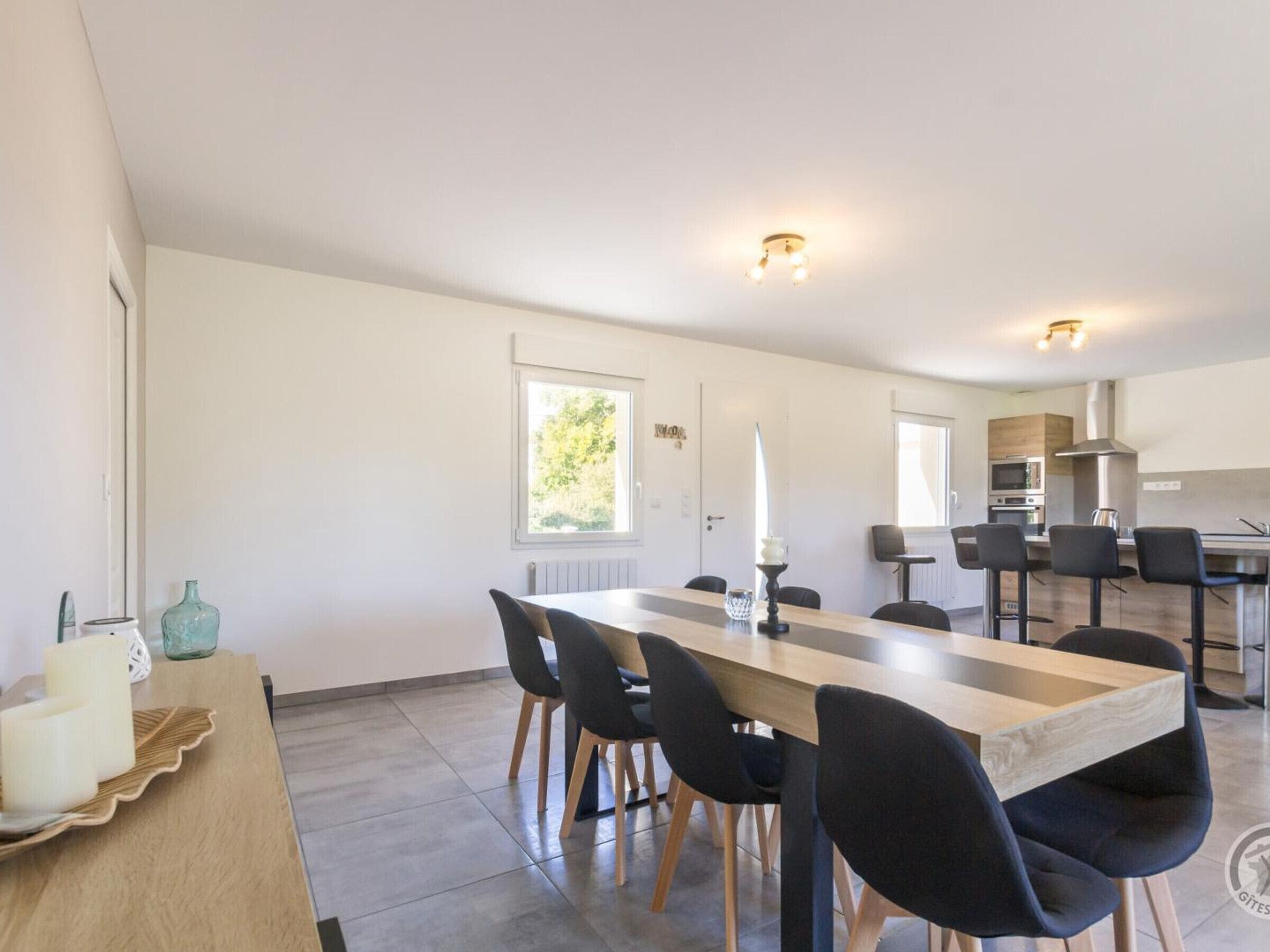 A modern kitchen with a central island, a dining area with a long table and chairs, and a view of the outdoors through a window.