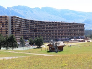 A large, multi-story hotel with a curved facade, surrounded by a grassy area and mountains in the background.