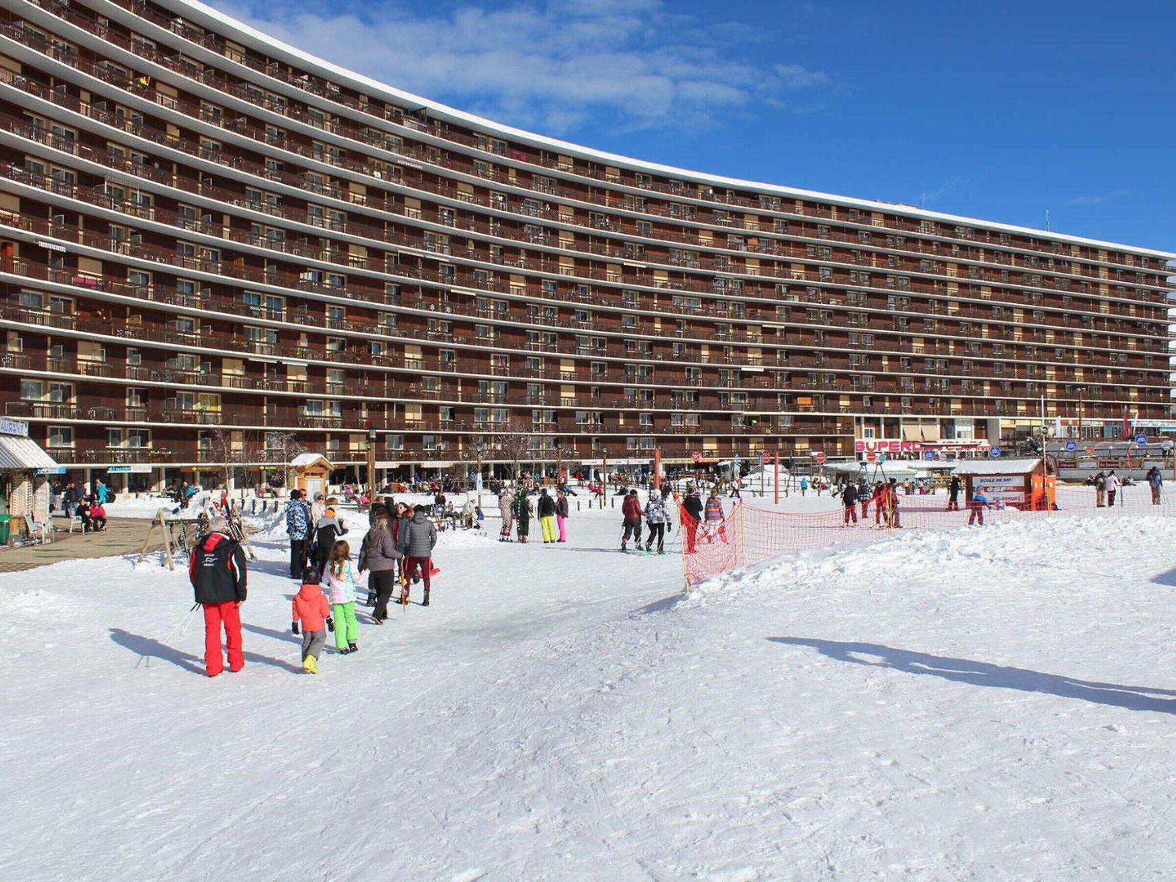 A large, multi-story hotel with numerous balconies, a ski resort area in the foreground, and a clear blue sky.