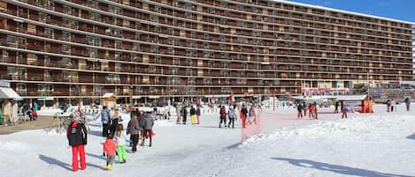 A large, multi-story hotel with numerous balconies, a ski resort area in the foreground, and a clear blue sky.