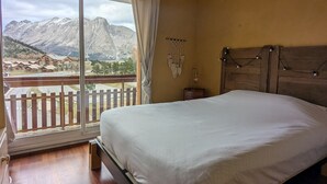 Un dormitorio con una cama grande, cabecera de madera, mesita de noche con lámpara y vistas a las montañas desde un balcón.