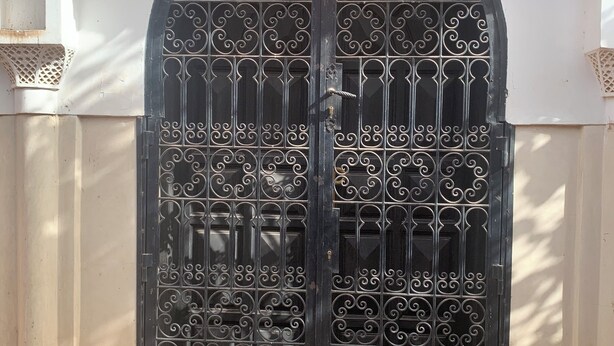 Una reja negra ornamentada con diseños intrincados, un banco de piedra en el frente y una pared blanca con detalles decorativos.