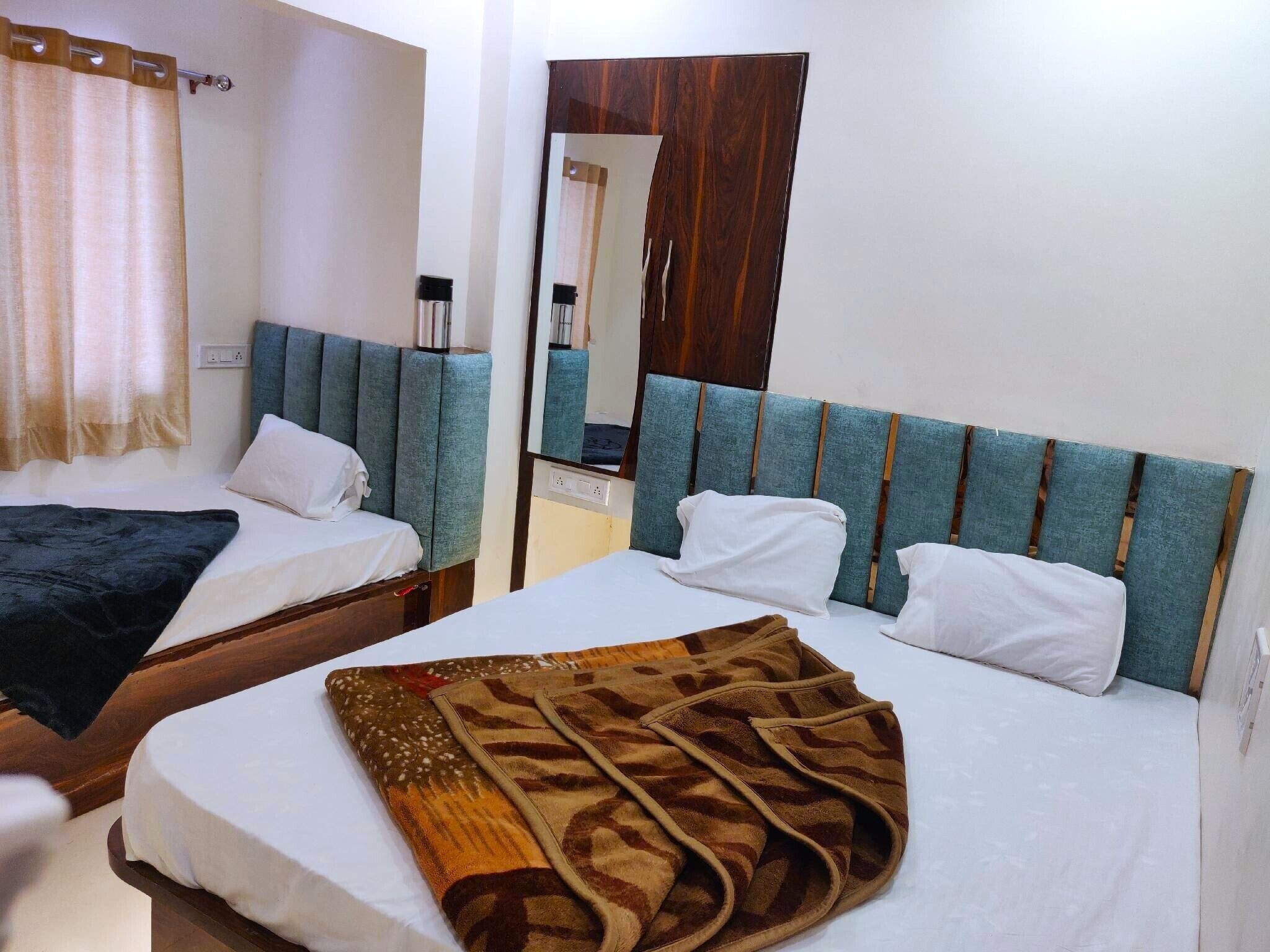 A hotel room with two beds, each with a headboard and pillows. There is a mirror on the wall and a window with curtains.