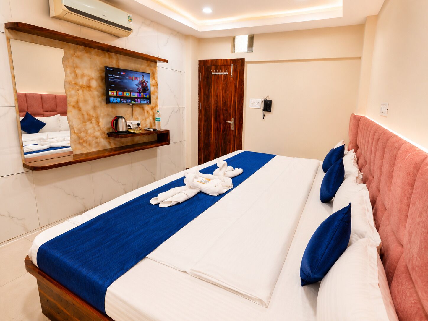 A hotel room with a bed, a TV mounted on a marble wall, a wooden door, and a bathroom visible in the background.