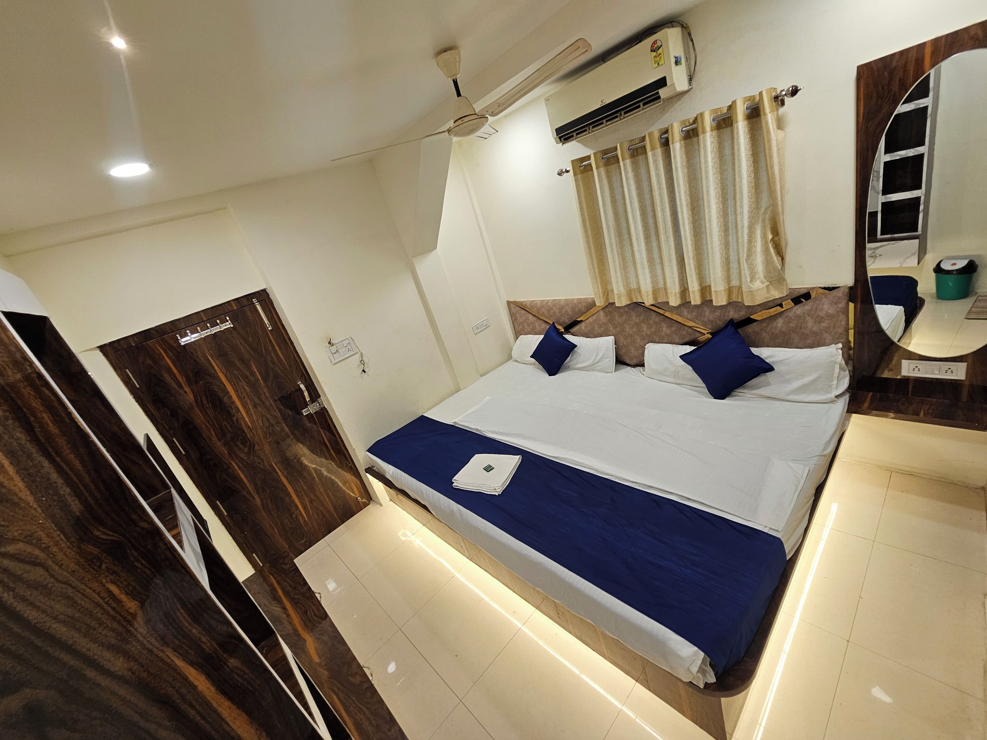 A hotel room with a bed, a ceiling fan, an air conditioning unit, a wooden door, a bathroom mirror, and a window with curtains.