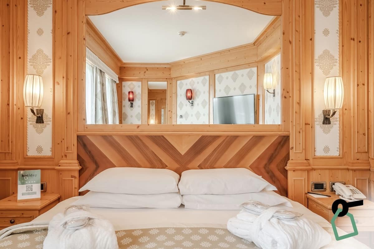 Une chambre d’hôtel avec une tête de lit en bois lambrissé, deux lits avec du linge de lit blanc, un miroir et une table de chevet avec un téléphone et un panneau indiquant « vacances ».