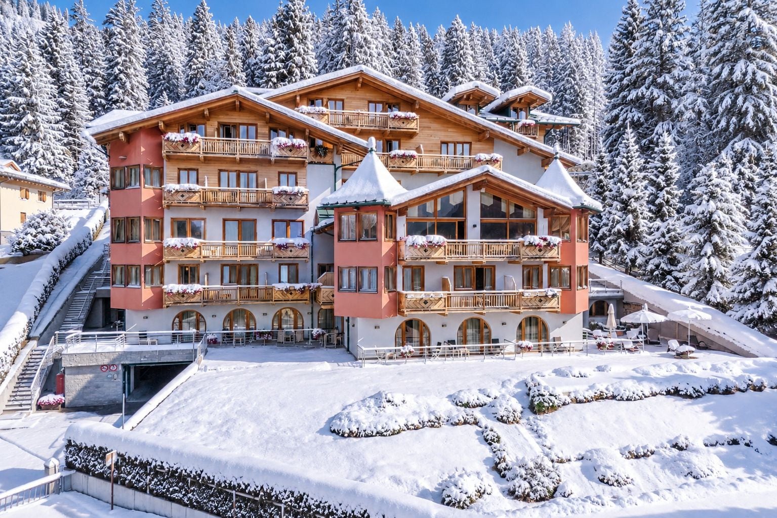 Un hotel a più piani con balconi, una pista da sci sullo sfondo e alberi innevati.