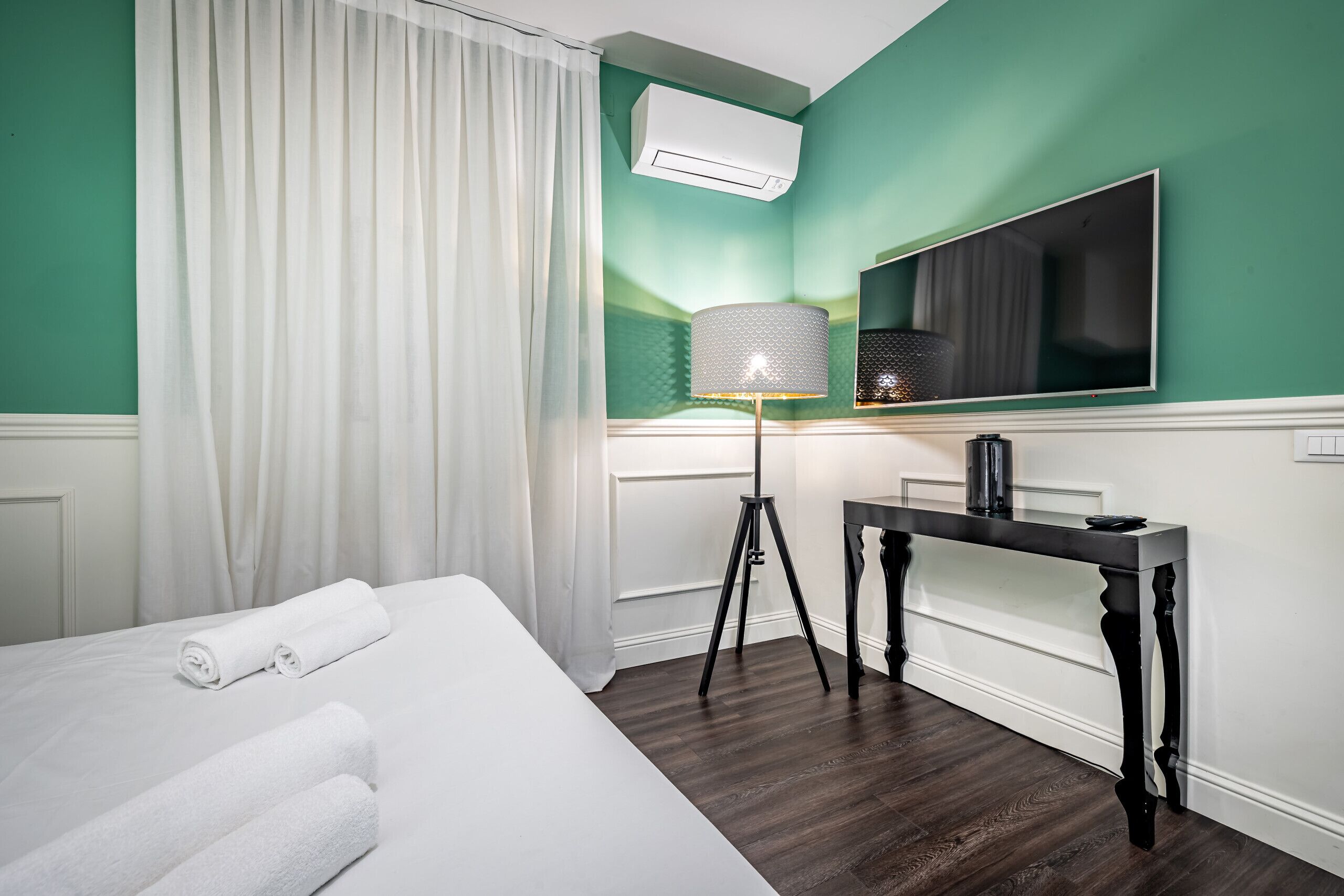 A hotel room with a bed, white curtains, a standing lamp, a flat-screen TV, and a dark wooden floor.