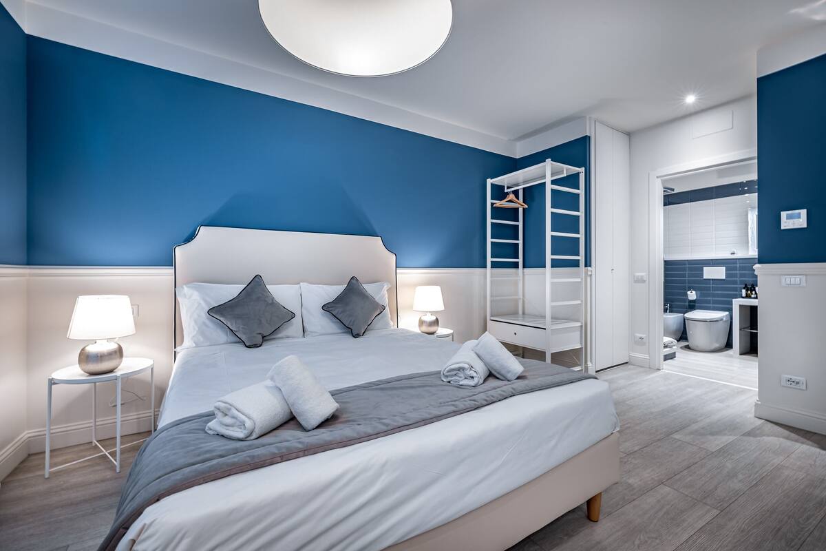 Une chambre moderne avec un grand lit, des lampes de chevet, une armoire et une salle de bain visible à travers une porte ouverte.