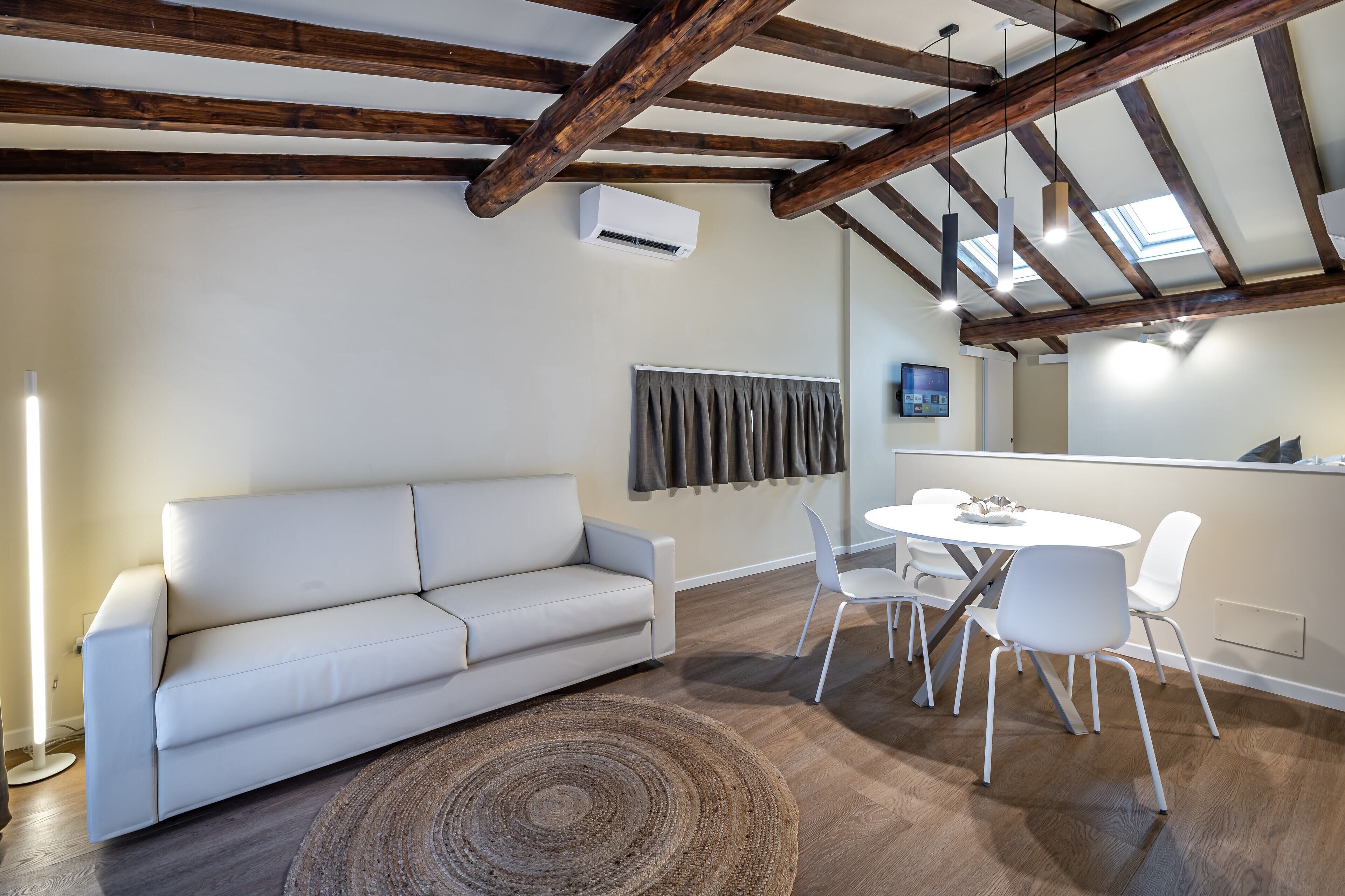 Ein modernes Wohnzimmer mit einem weißen Sofa, einem runden Couchtisch und weißen Sesseln. Die Holzbalkendecke ist sichtbar und an der Wand ist eine Klimaanlage angebracht.