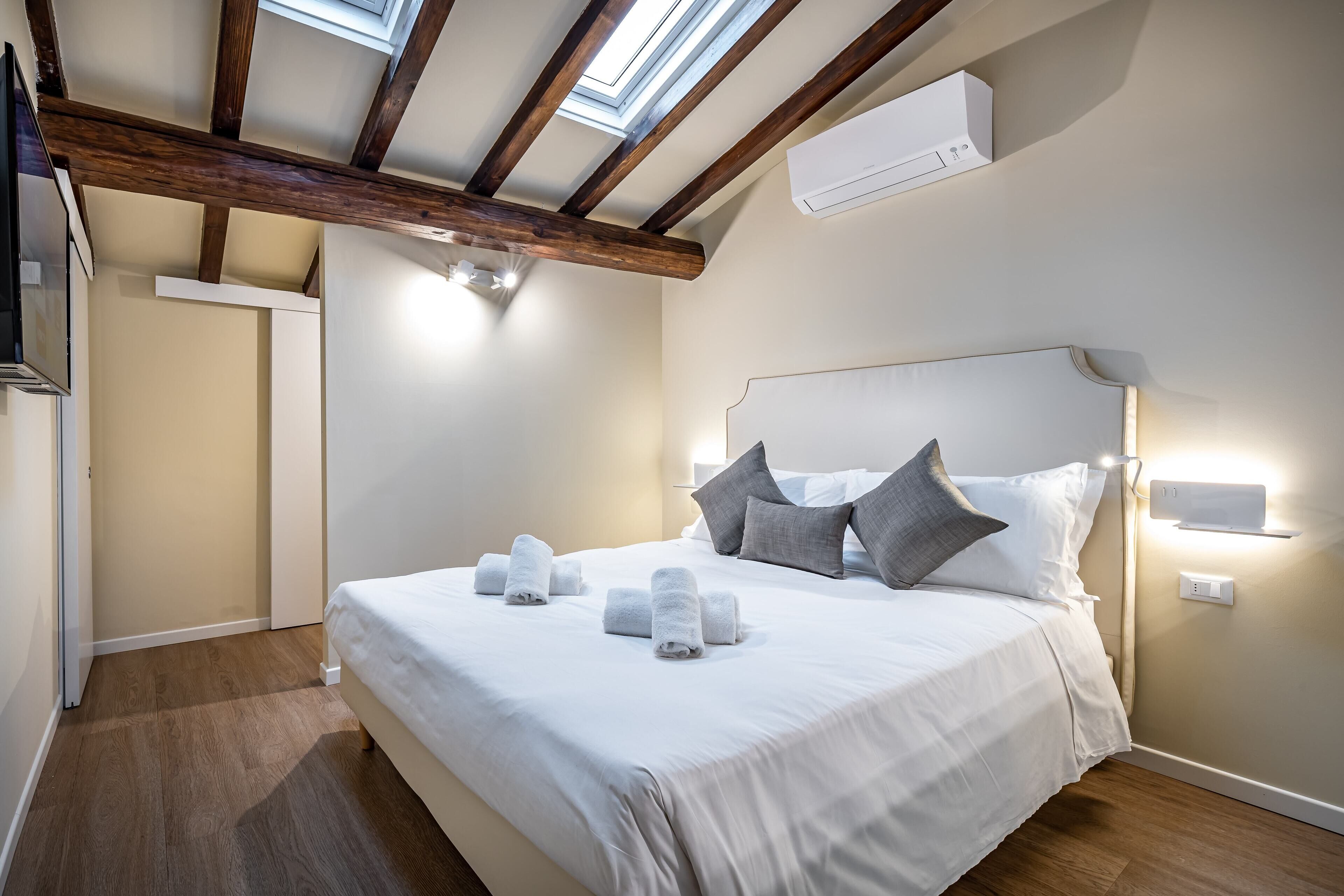 A bedroom with a wooden ceiling, a bed with white bedding and towels, two bedside lamps, and an air conditioning unit.