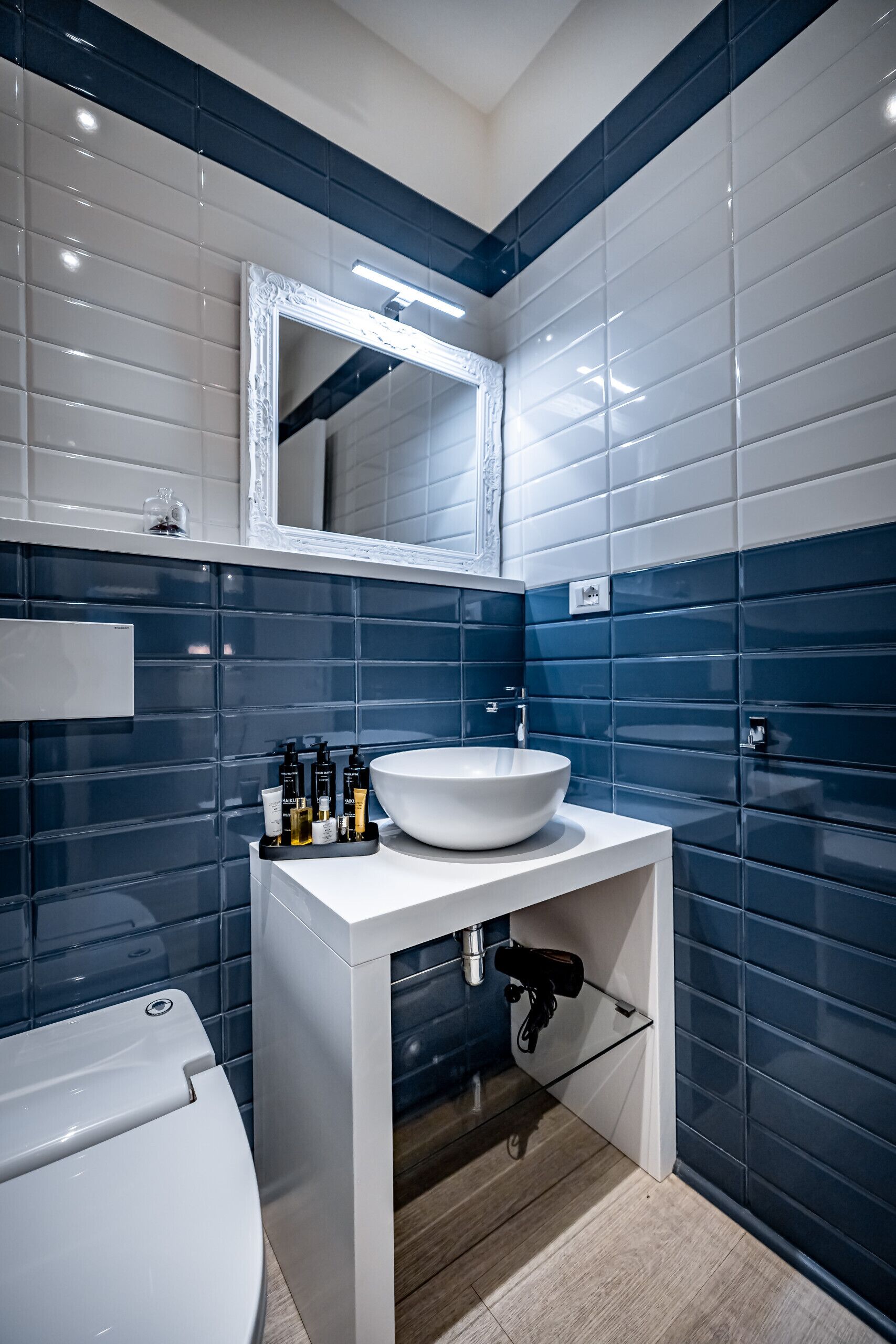 Un baño moderno con un lavamanos blanco, paredes con azulejos azules, un espejo sobre el lavamanos y un inodoro a la izquierda.