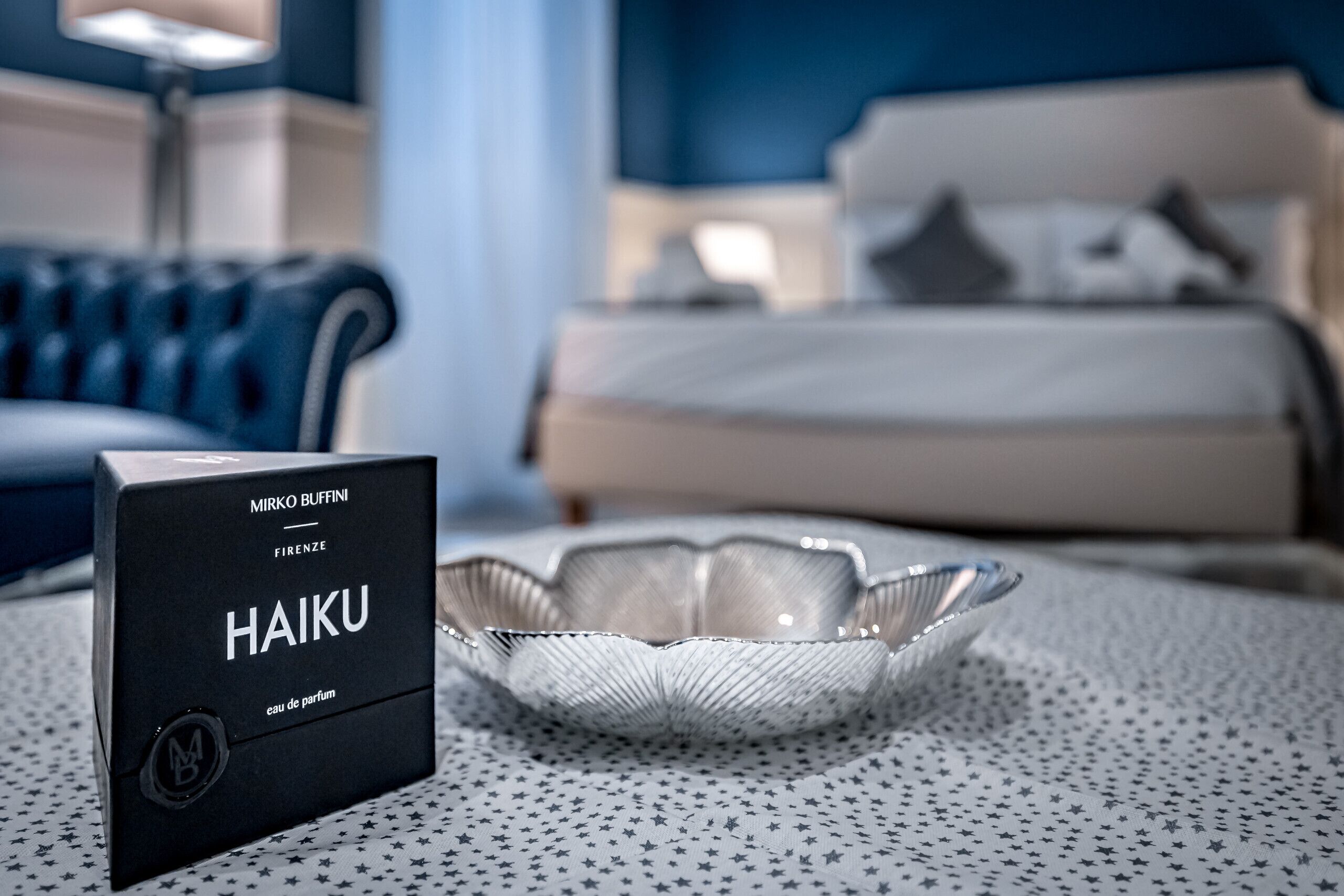 Ein Hotelzimmer mit einem ordentlich bezogenen Bett, einem Teppich mit Sternenmuster und einer Parfumschachtel mit dem Aufdruck 'HAIKU' auf dem Boden.