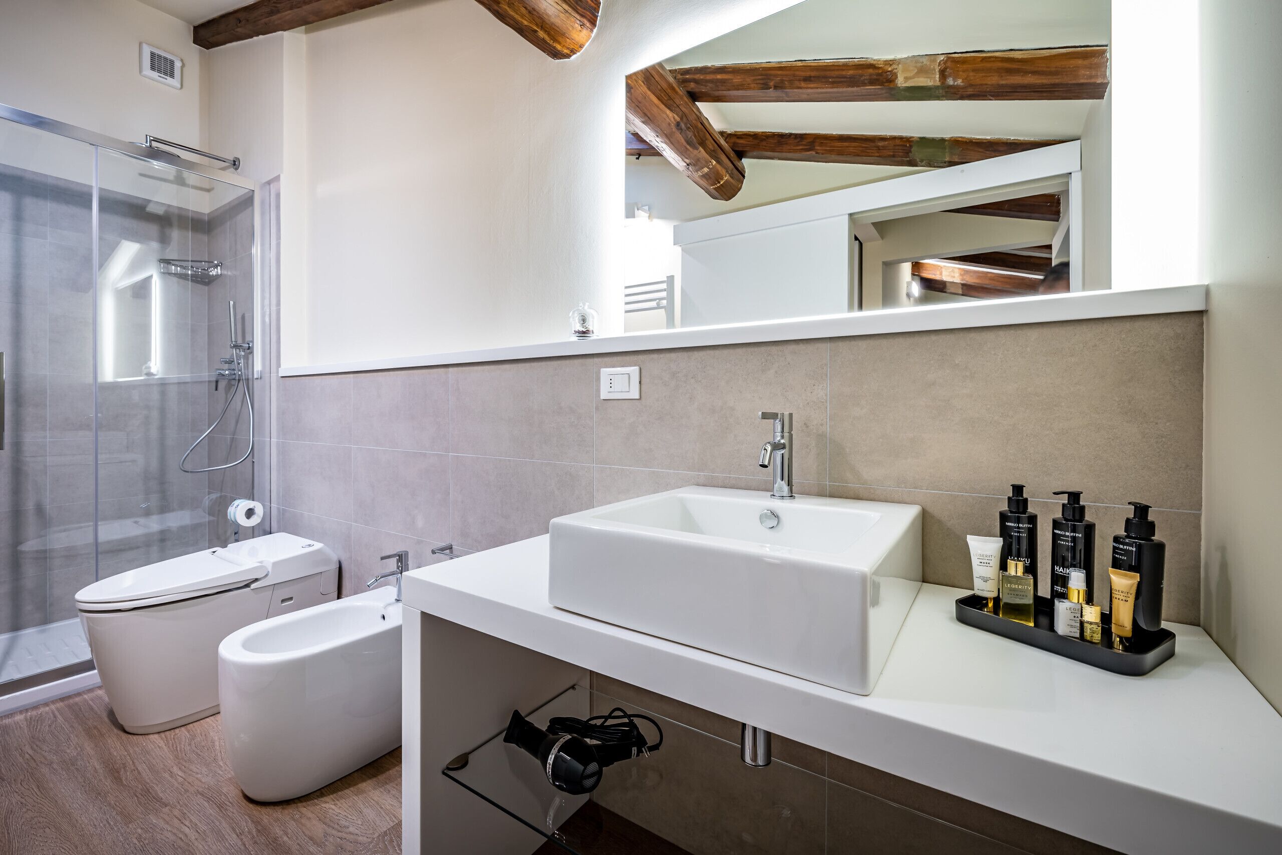 A modern bathroom with a glass shower enclosure, a bidet, a sink with toiletries, and a mirror above the sink.