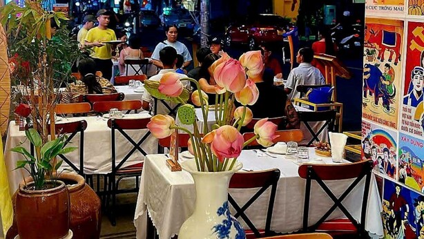 Uma área de refeições ao ar livre com mesas, cadeiras e um vaso grande com flores.