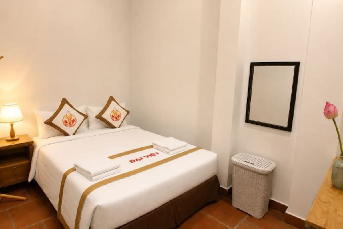 Quarto de hotel com uma cama bem arrumada, uma lixeira, uma mesinha de cabeceira com abajur e um quadro na parede.