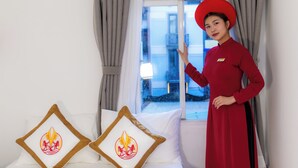 Quarto de hotel com uma cama, duas almofadas com um logotipo, uma janela e uma pessoa de uniforme vermelho.