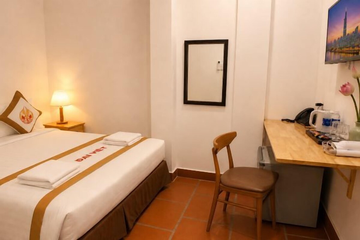 A hotel room with a bed, a desk, a chair, a lamp, and a framed picture on the wall.