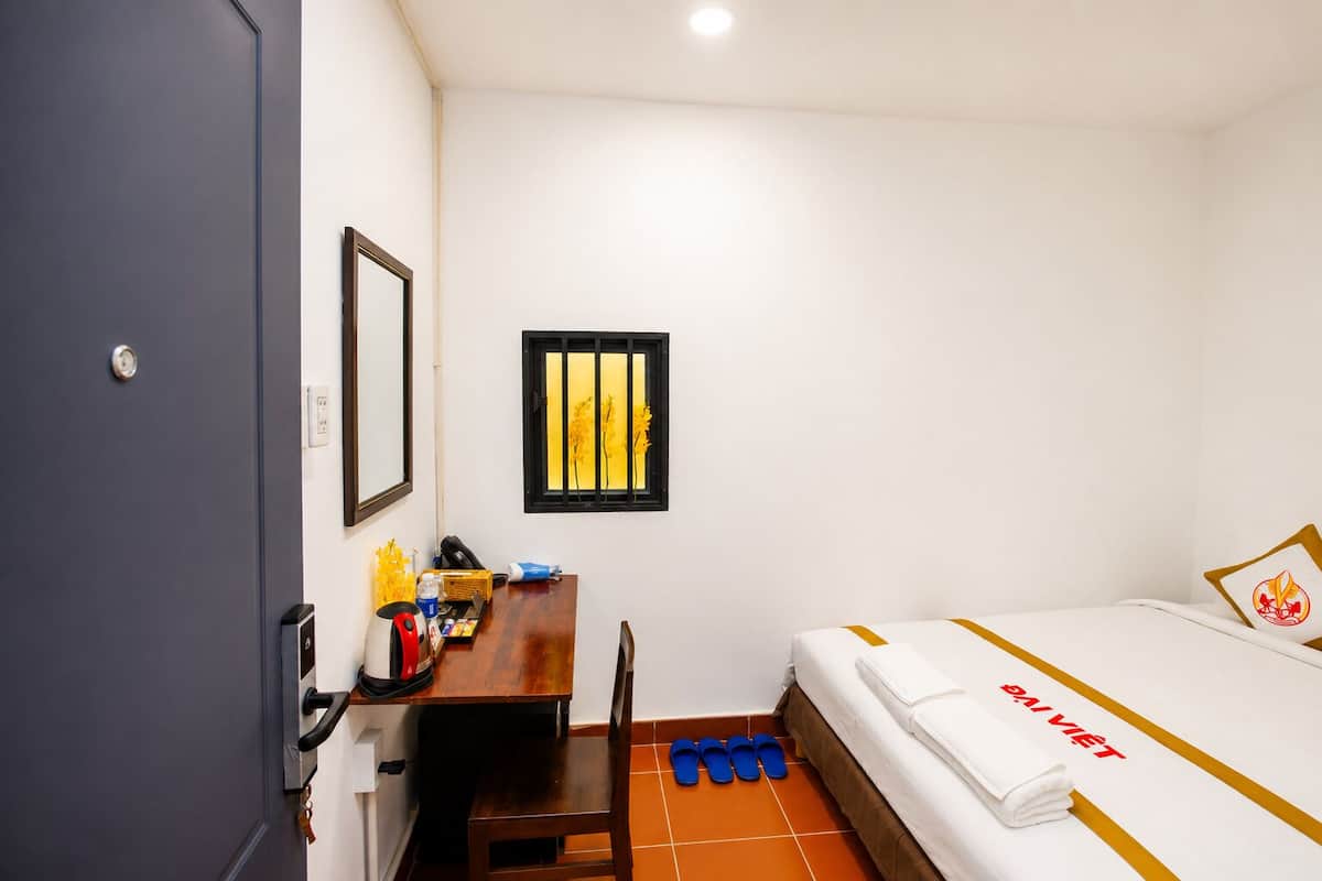 A hotel room with a bed, a desk, a chair, a window with yellow curtains, and a door.