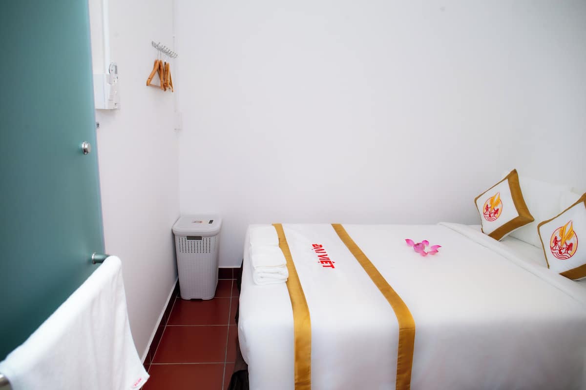 A hotel room with a bed, a white wall, a green door, a white trash bin, and a white towel hanging on the door.