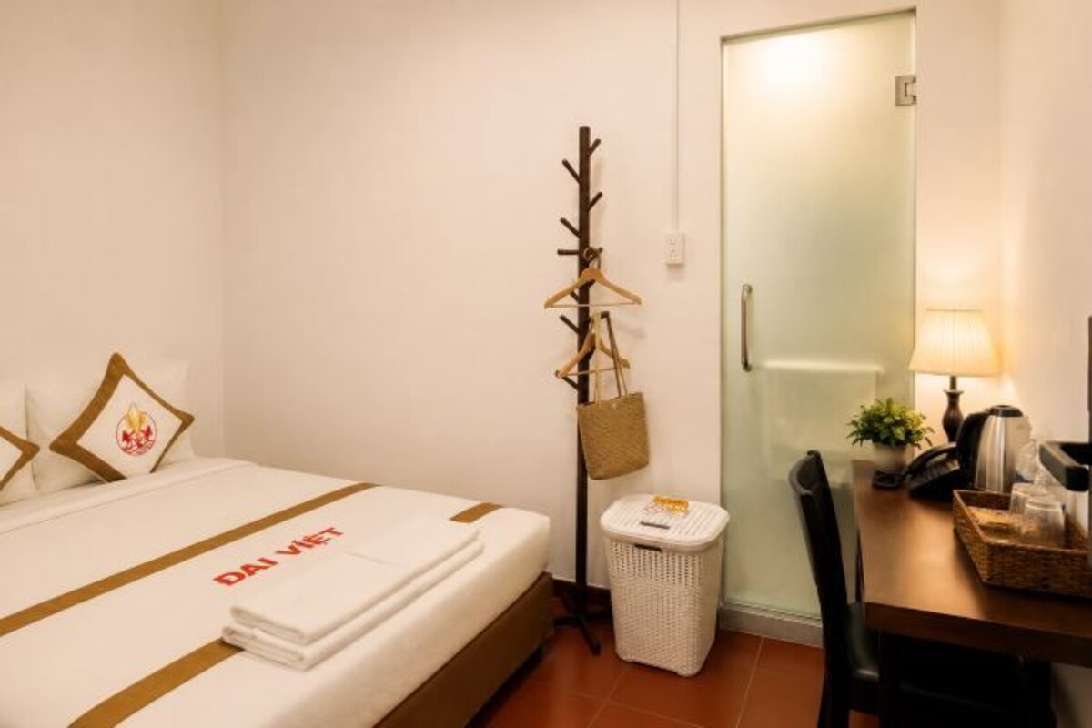 Quarto de hotel com cama, escrivaninha, cadeira, abajur e lixeira pequena.