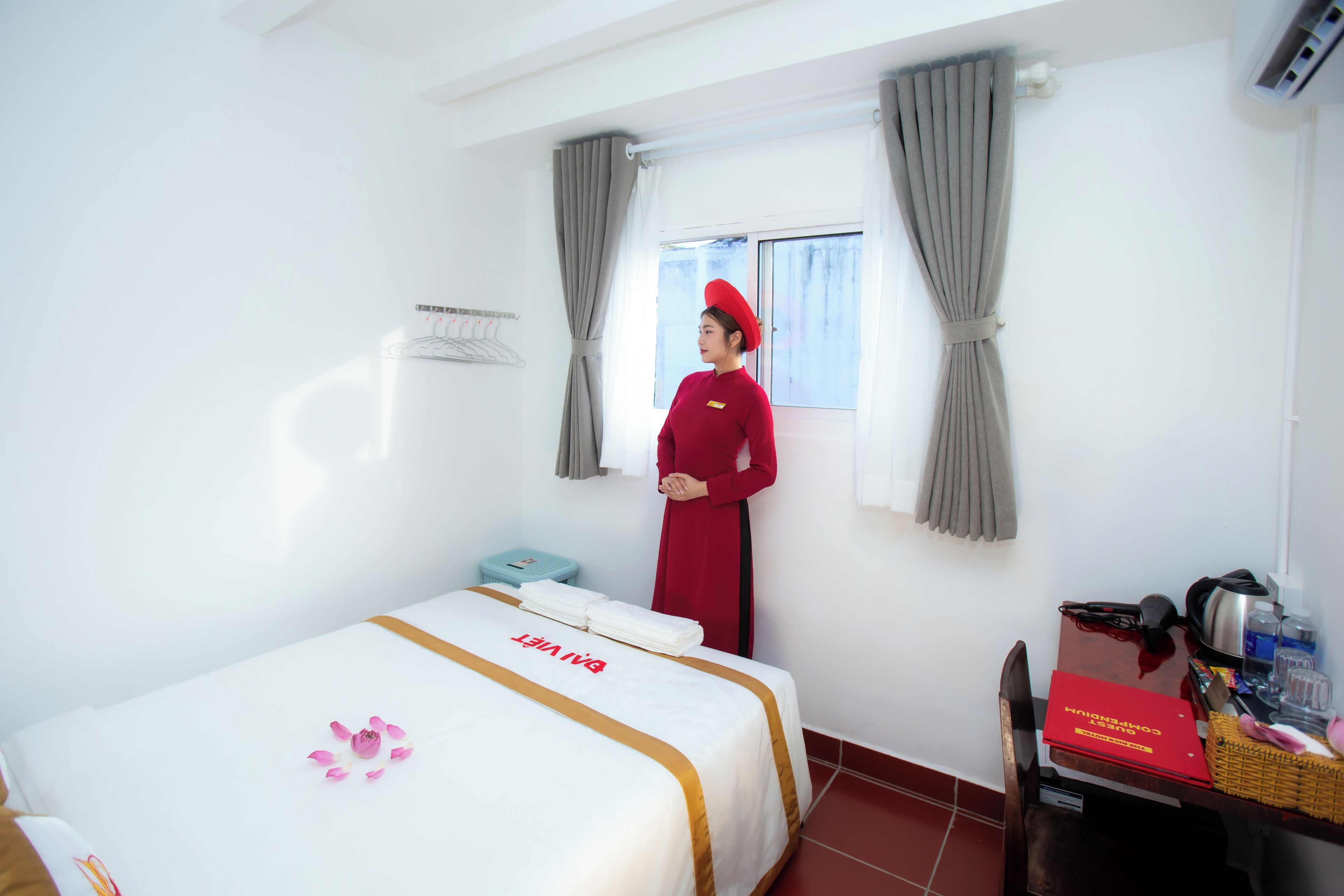 Habitación de hotel con cama, ventana con cortinas, escritorio y una caja roja, y una persona con uniforme rojo.